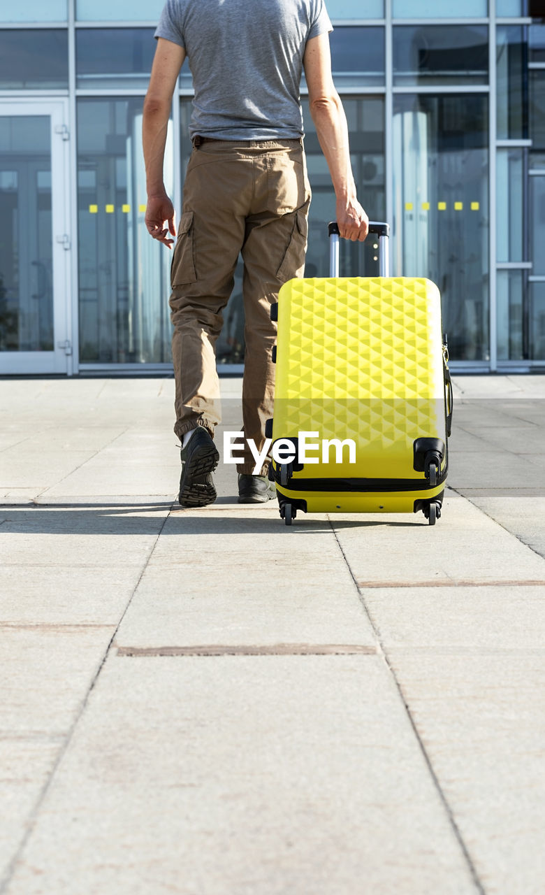 Man traveler in casual carrying yellow suitcase next to entrance to airport outside  vacation mockup