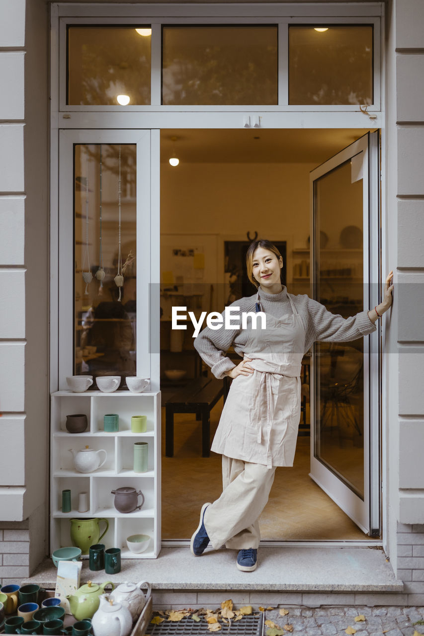 Smiling young female potter standing with hand on hip at workshop doorway