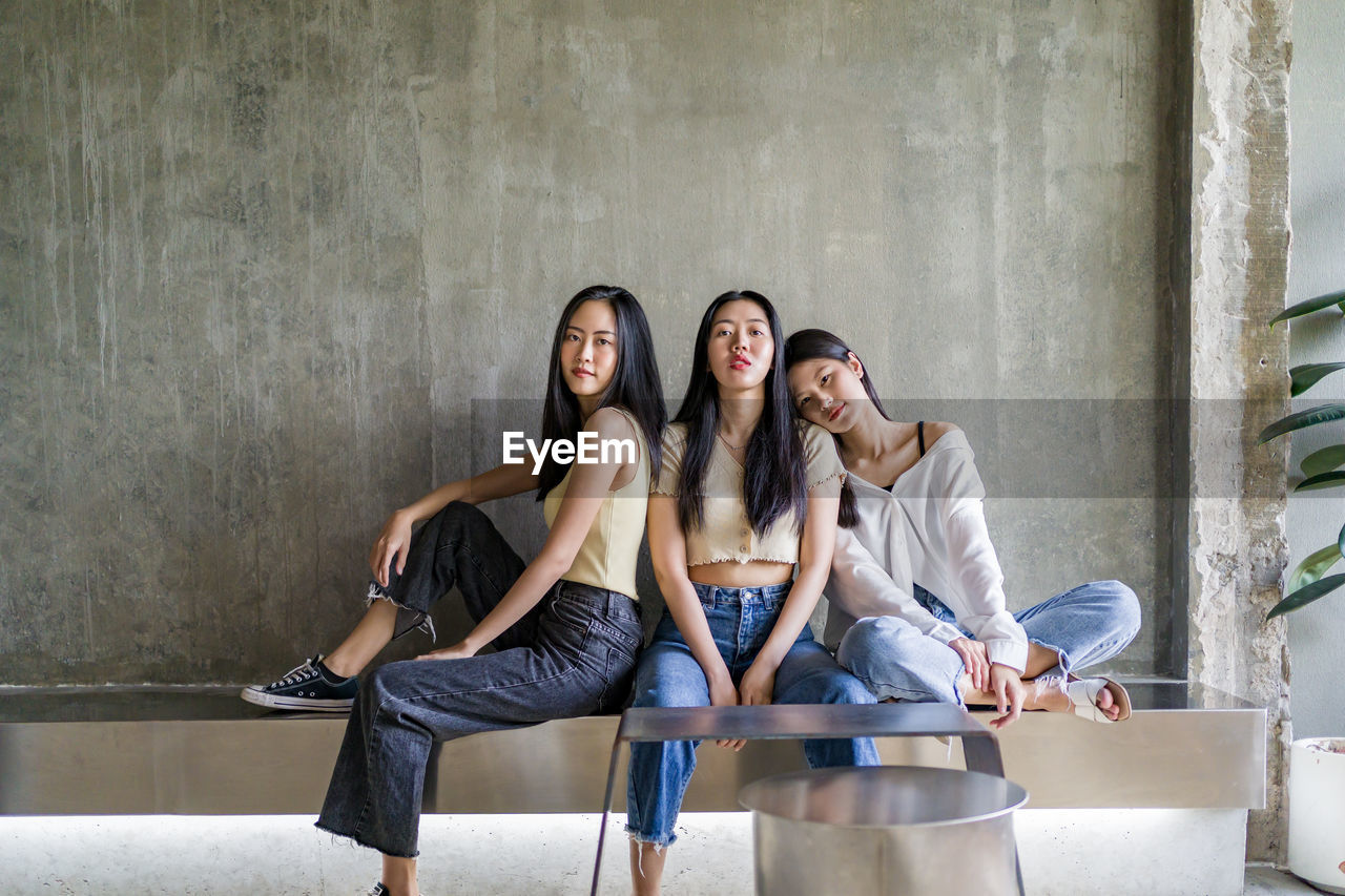 Portrait of smiling young women sitting on bench against wall