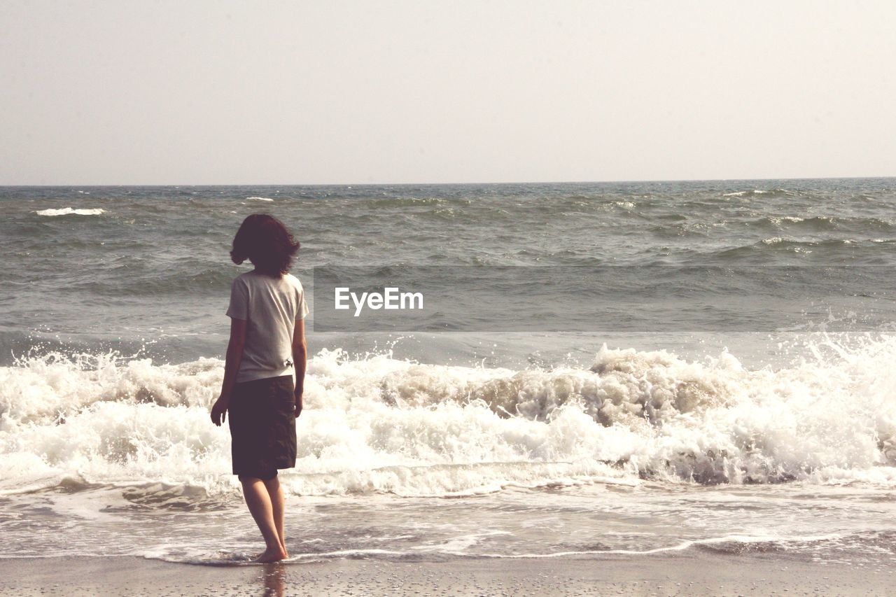 Rear view of boy standing on beach against clear sky