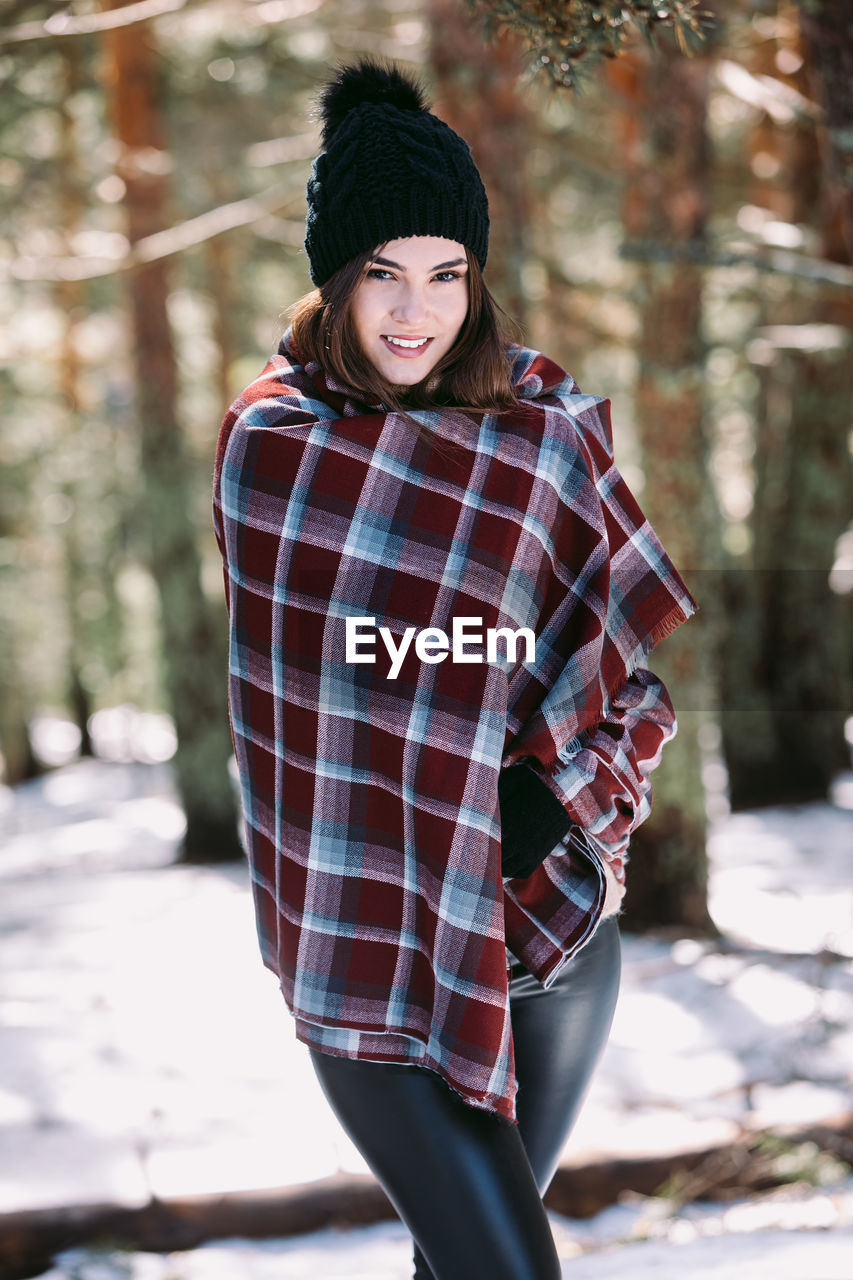 Content female wrapped in warm plaid standing in snowy woods and looking at camera