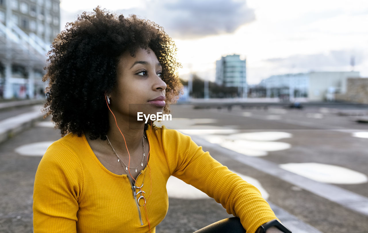 Thoughtful woman with afro hair listening music while sitting against sky in city