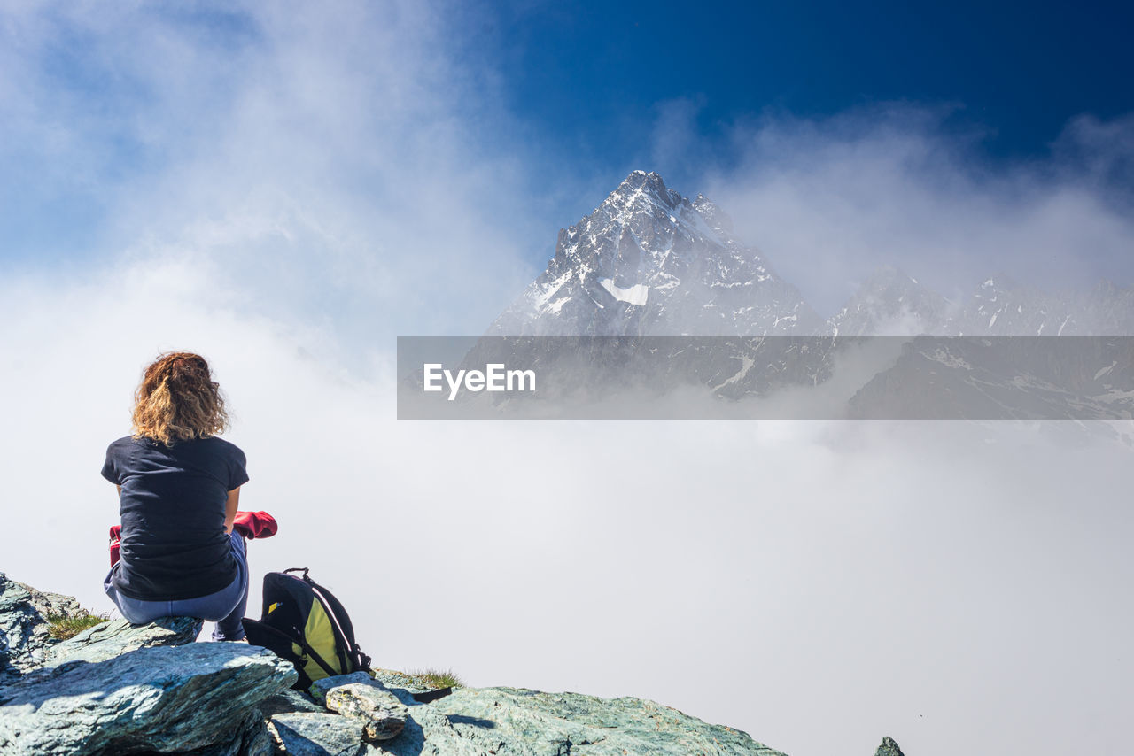 REAR VIEW OF WOMAN SITTING ON ROCKS AGAINST MOUNTAIN