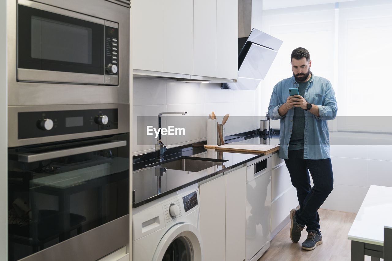 Man cooking crepes in the kitchen with a mobile phone in a denim shirt