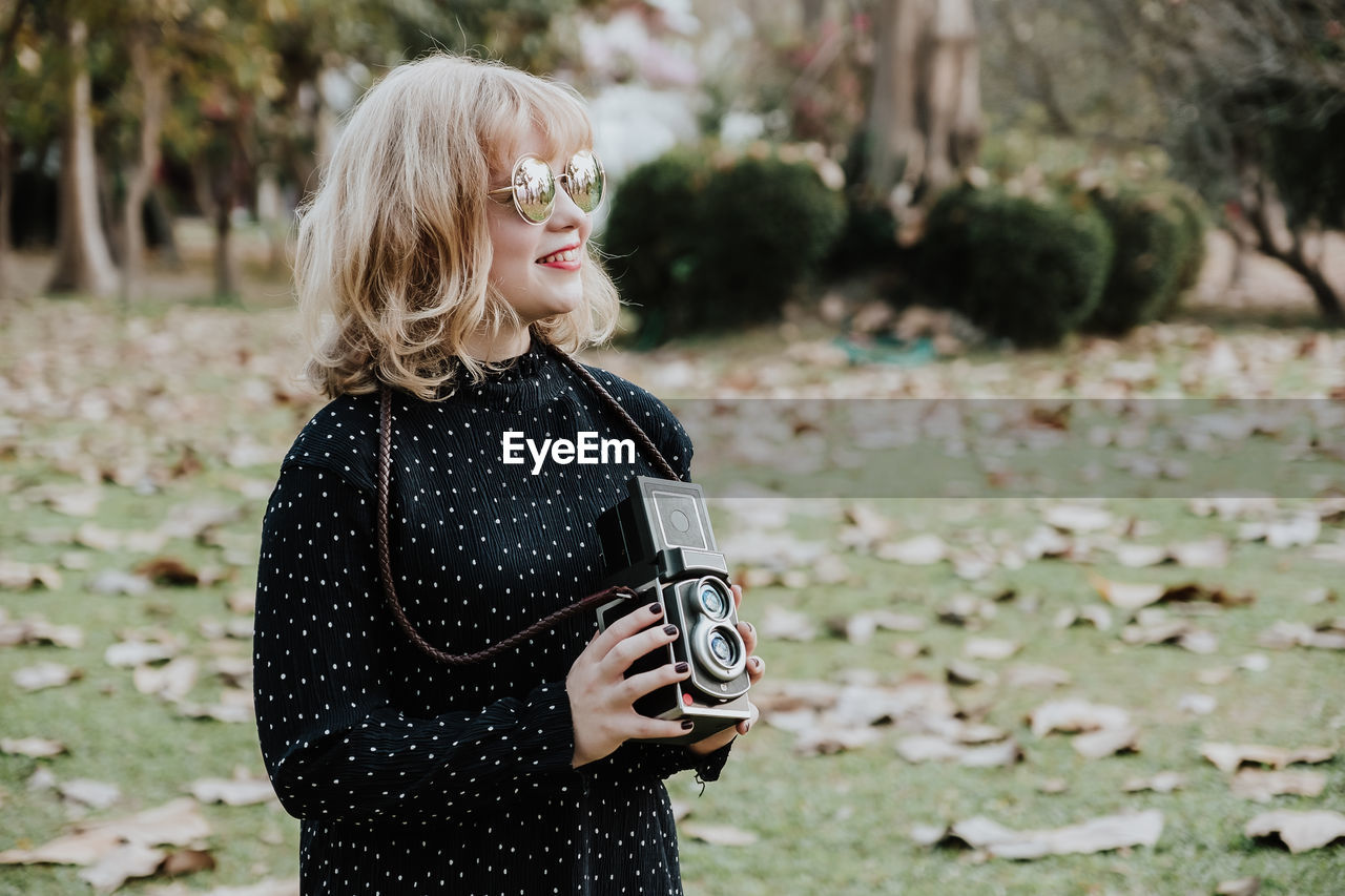 Smiling young woman holding vintage camera while standing on land during autumn