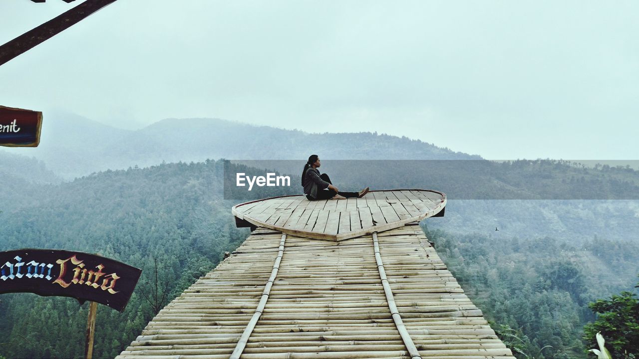 Side view of woman sitting on wood against sky