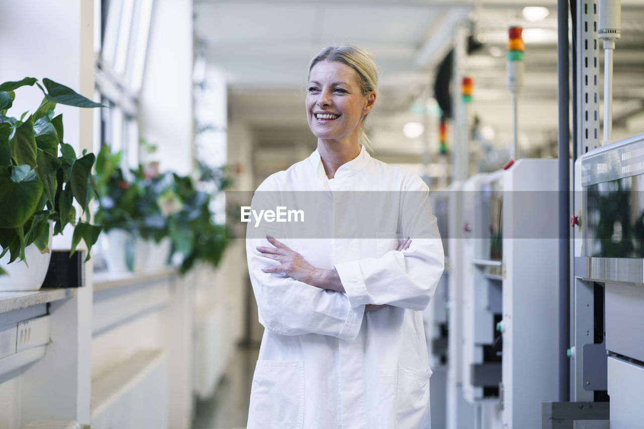 Confident mature female scientist with hands in lab coat pockets standing by machinery at laboratory