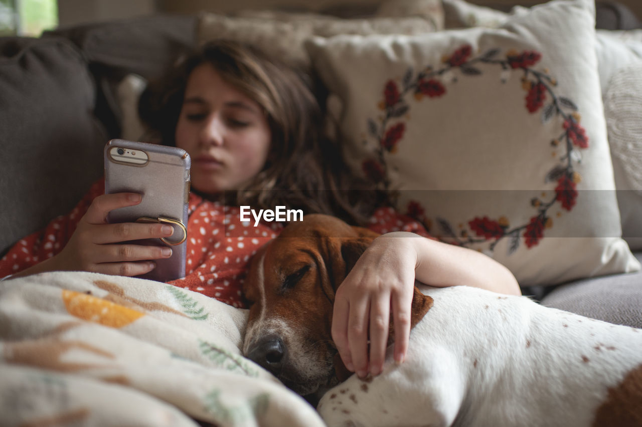 Tween girl looking at her phone while cuddling with hound dog on couch
