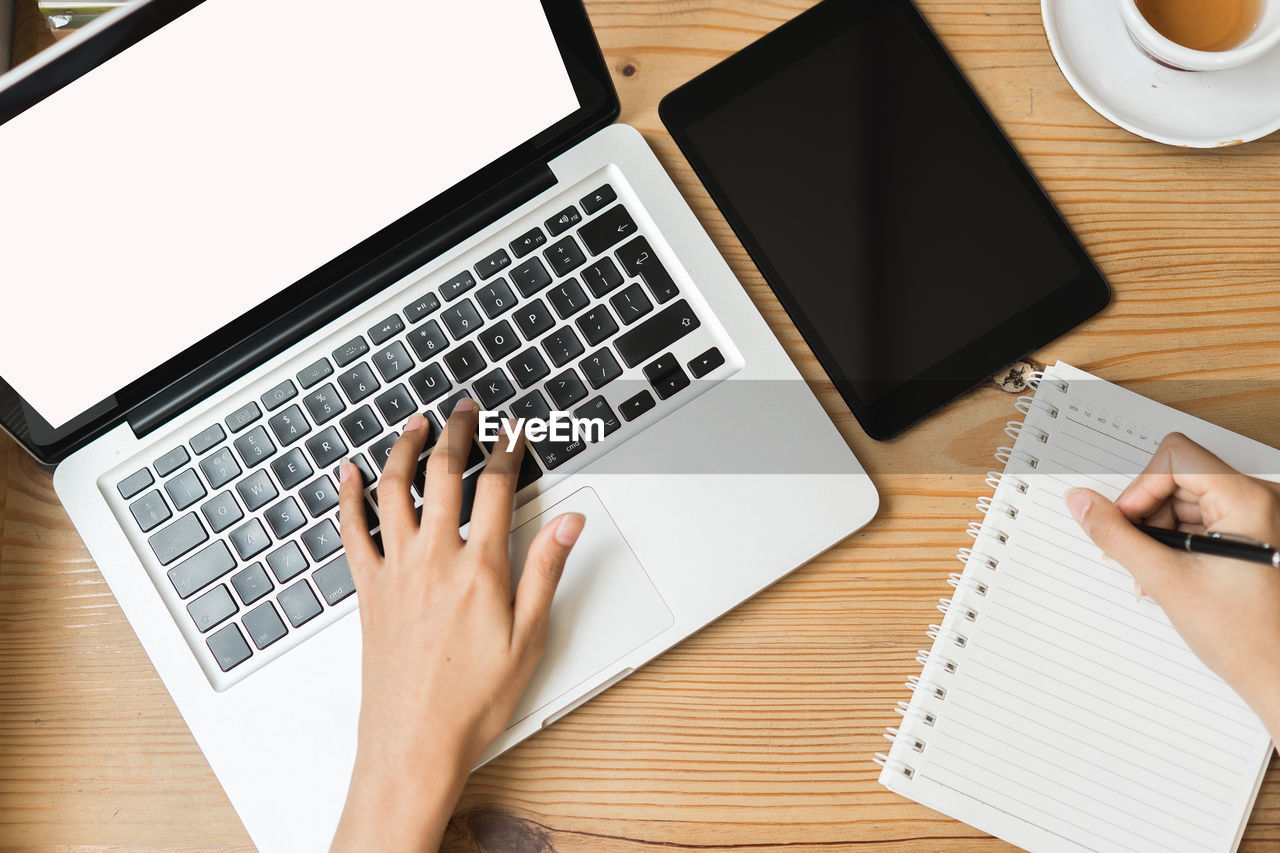 Cropped hands of businessman writing on book while using laptop at desk