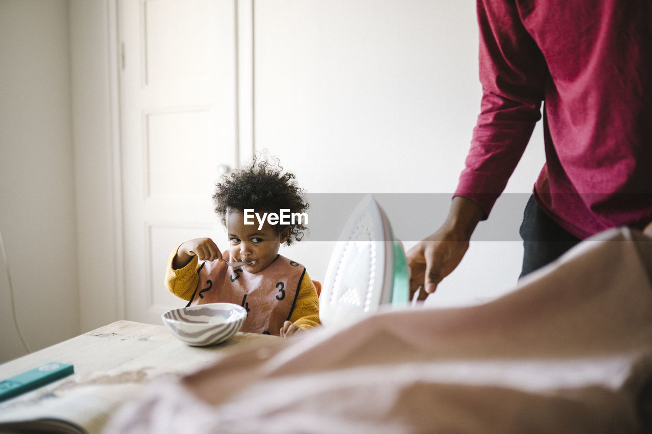 Girl eating from bowl while father ironing clothes