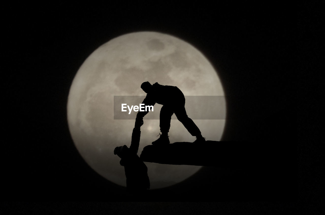 Silhouette person helping friend to climb on rock against moon at night