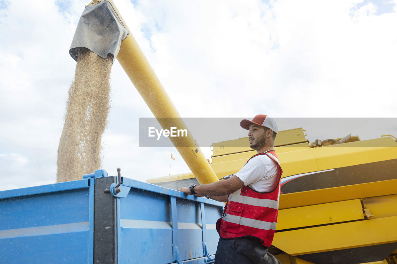 Side view of male farmer standing near industrial combine harvester unloading dried grains into trailer while working in agricultural plantation