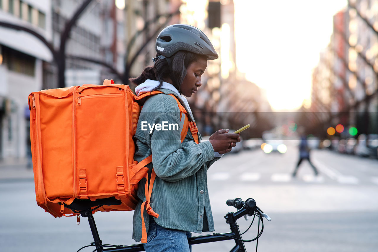 Side view of african american female cyclist with thermo backpack standing on paved sidewalk while checking order information on cellphone