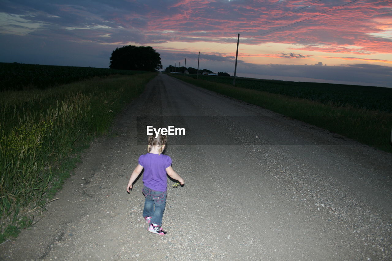 Rear view full length of girl standing on road amidst grassy field during sunset