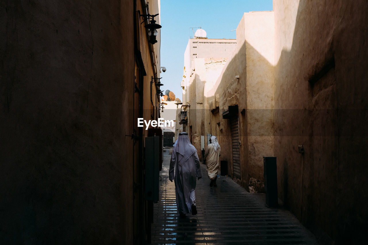 Rear view of men wearing traditional clothing walking amidst buildings at alley