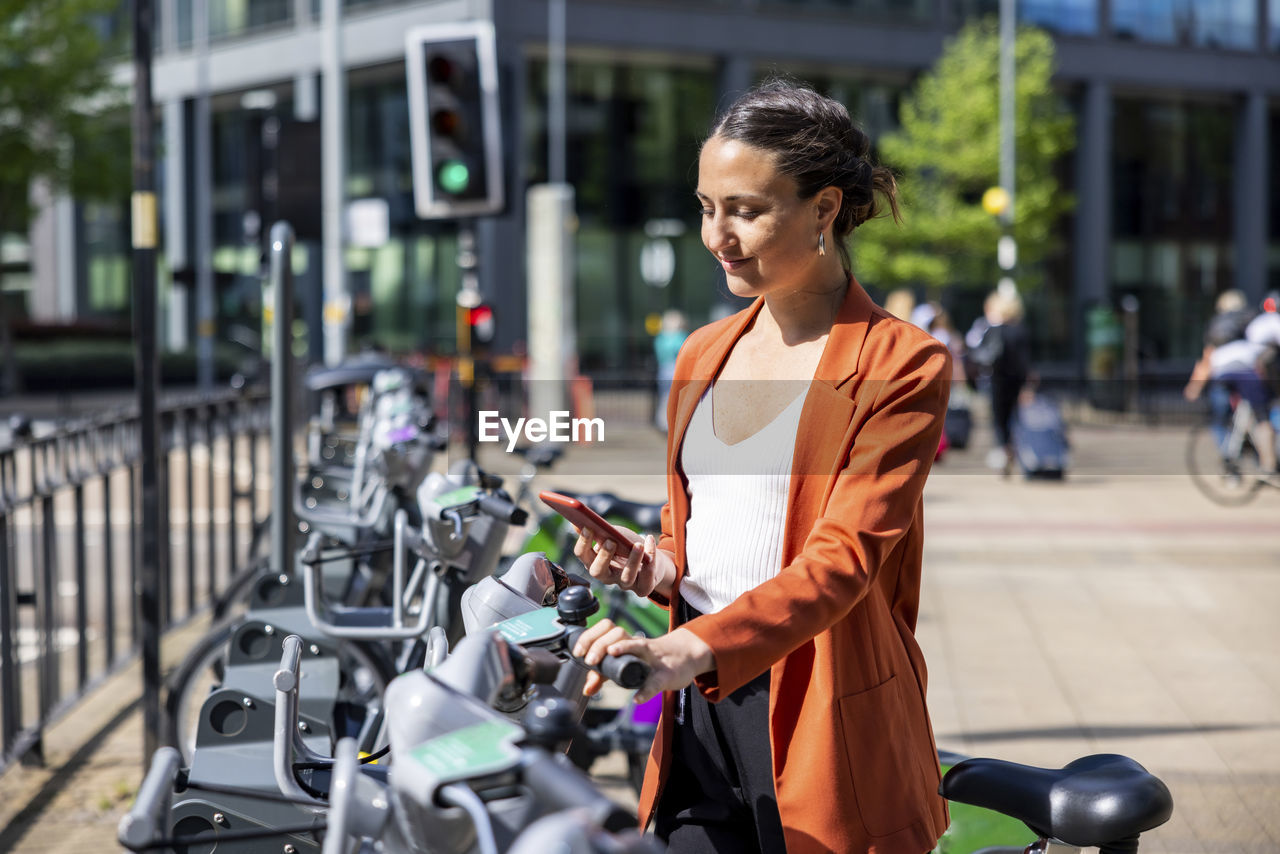 Smiling businesswoman holding mobile phone standing by electric bicycle