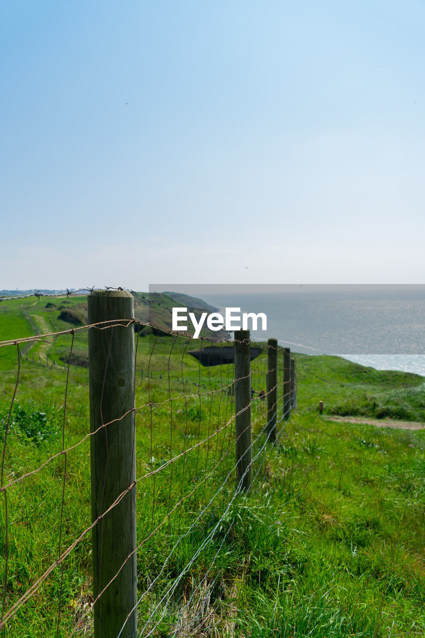 land, sky, fence, sea, grass, water, scenics - nature, nature, horizon, plant, security, tranquility, landscape, tranquil scene, protection, beauty in nature, coast, horizon over water, no people, beach, environment, green, hill, field, clear sky, wire, day, non-urban scene, barbed wire, cliff, outdoors, rural scene, idyllic, terrain, coastline, travel destinations, blue, rural area, travel, tourism, copy space, wood, architecture