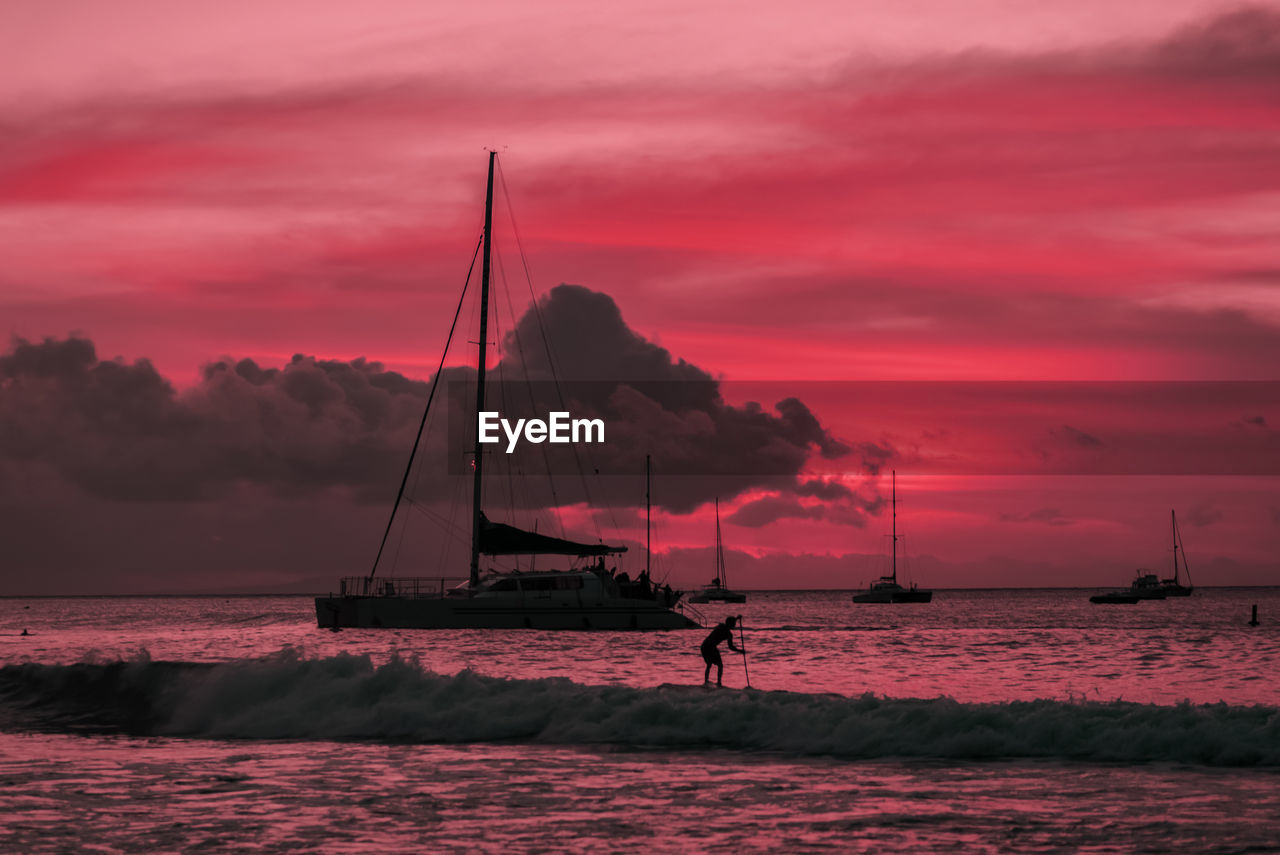 Silhouette man paddleboarding by boats on sea against dramatic sky