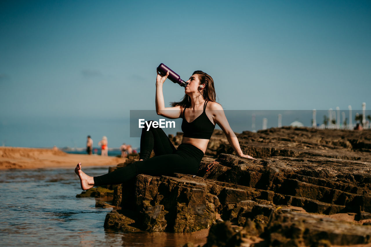 Peaceful female in sportswear sitting on stone near river drinking water while doing yoga
