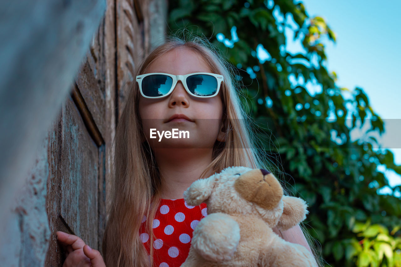 Beautiful happy girl in red polka dot dress soft toy on wooden balcony. cute joyful child long hair.