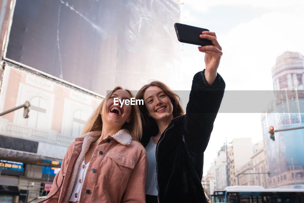 Low angle of cheerful female friends standing on street in madrid and taking self shot on smartphone during city stroll