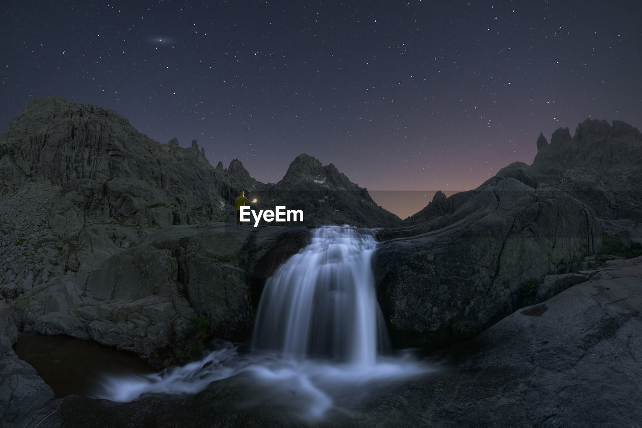 Traveler admiring cascade with foam on rough mount against pond under starry sky at dusk