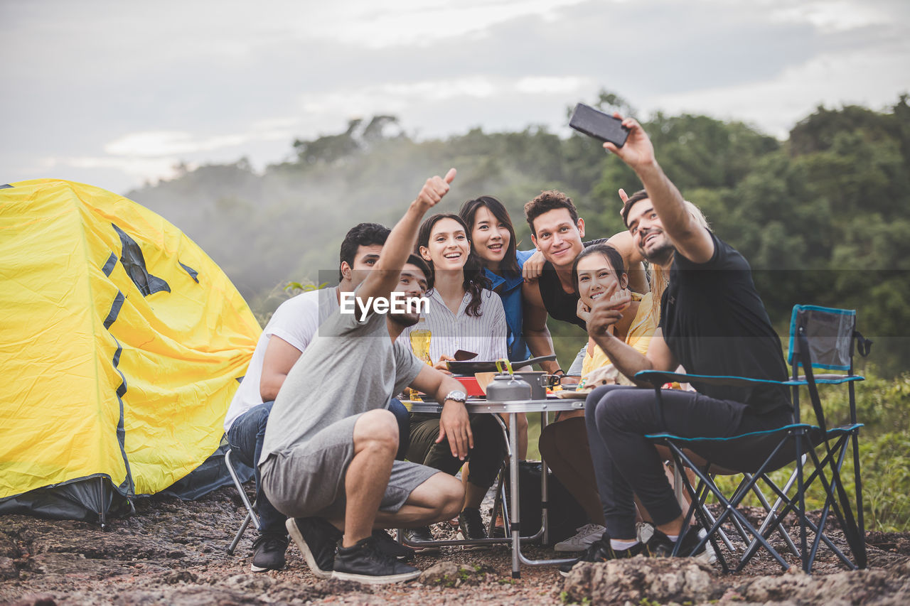Friends taking selfie while sitting on land by tent against sky