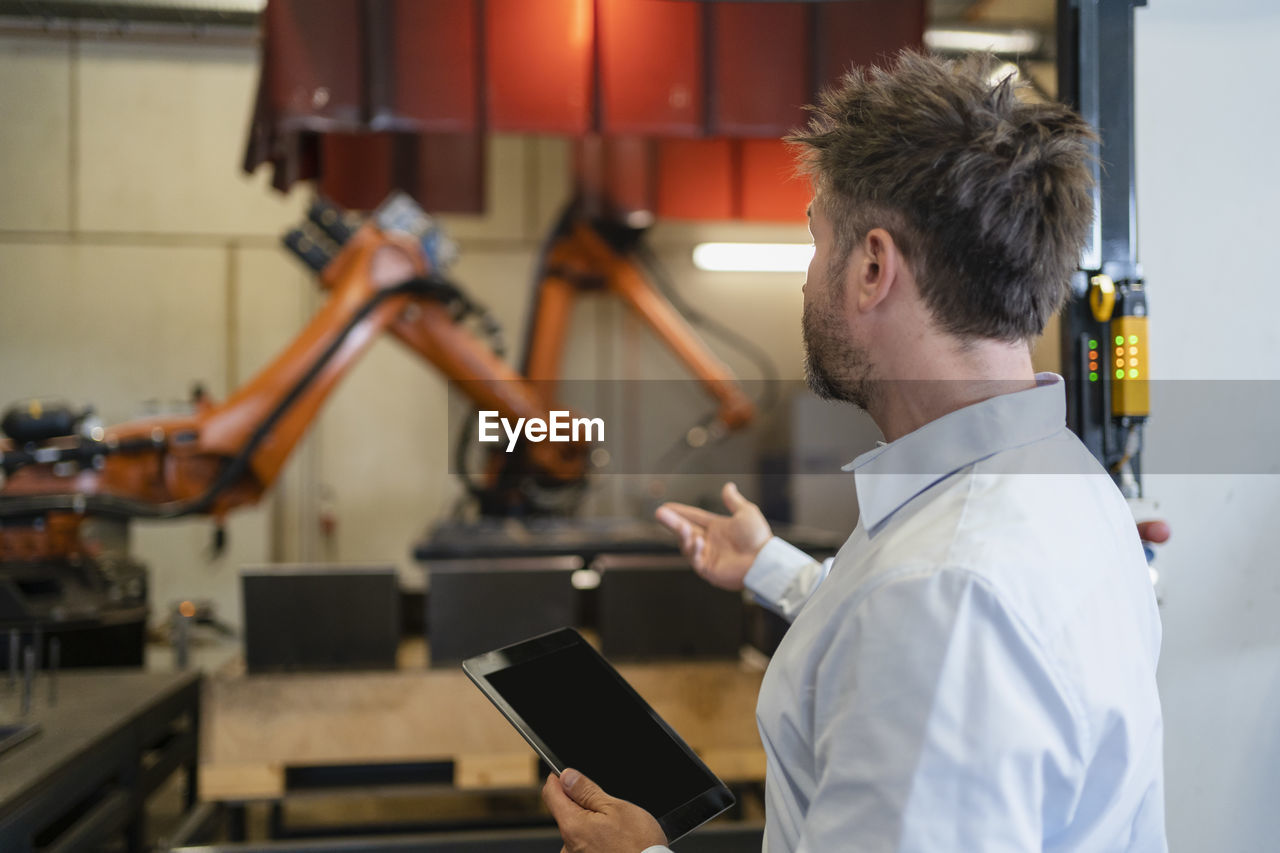 Mature businessman with digital tablet gesturing toward robotic arm machine while standing at factory