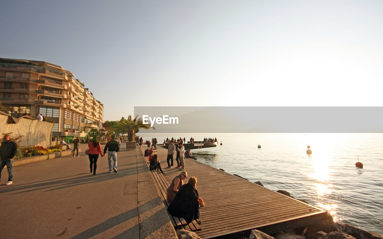 People at shore by buildings against clear sky