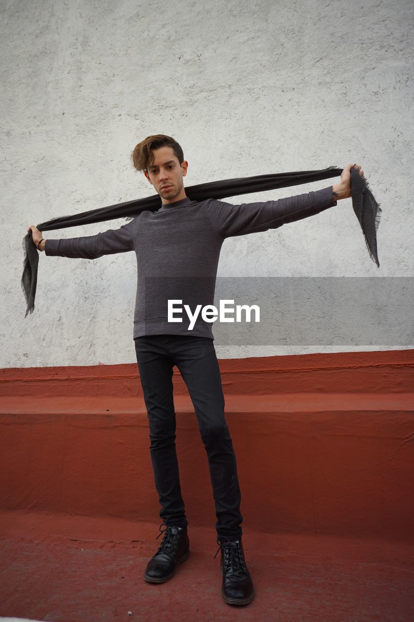 Full length of young man holding scarf while standing against wall