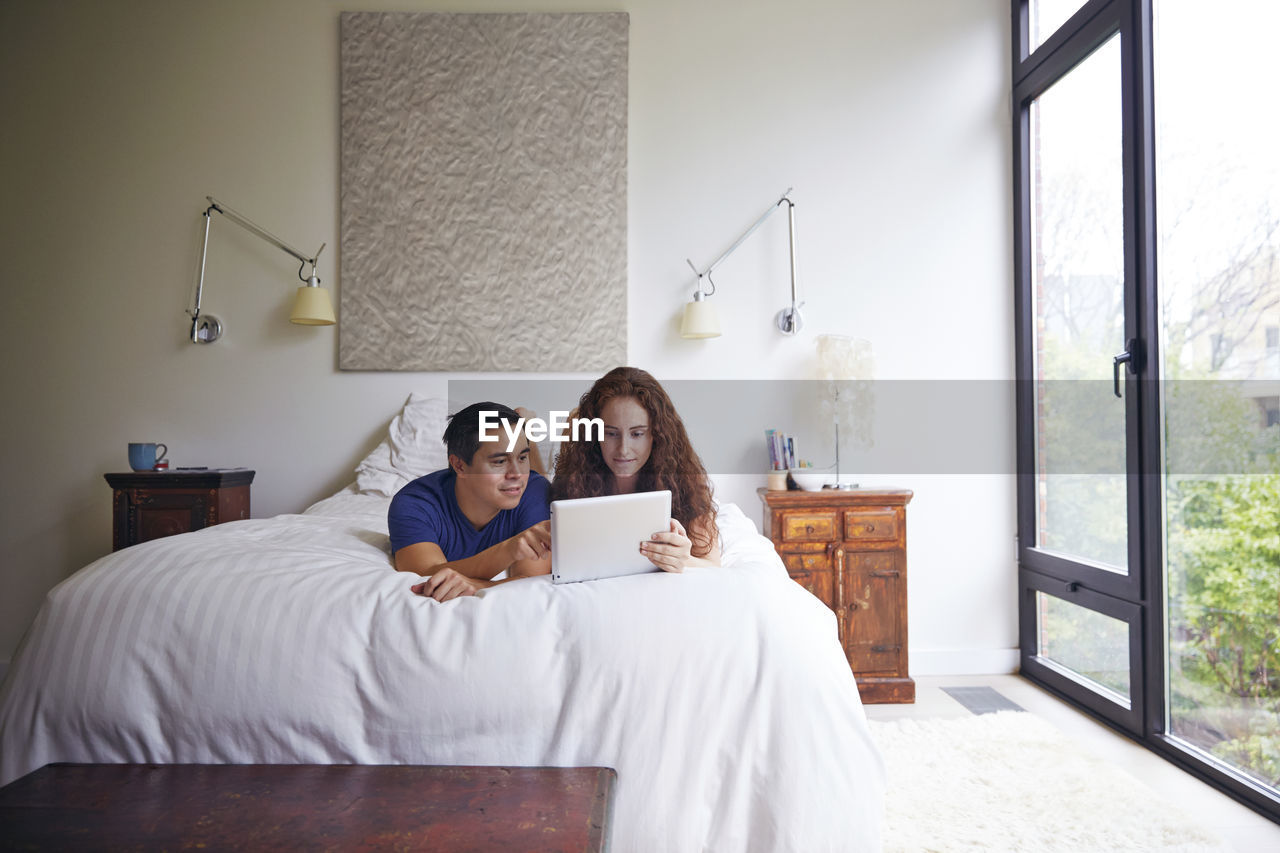 Couple looking at tablet computer while lying on bed against wall
