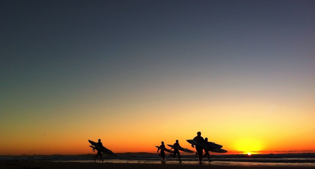 Silhouette people with surfboards running on beach at sunset