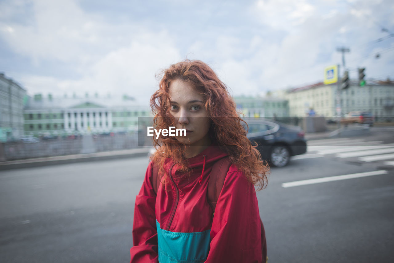 Serious millennial redhead female with long curly hair wearing red jacket looking at camera while standing alone against road and historic buildings in windy day in saint petersburg in russia