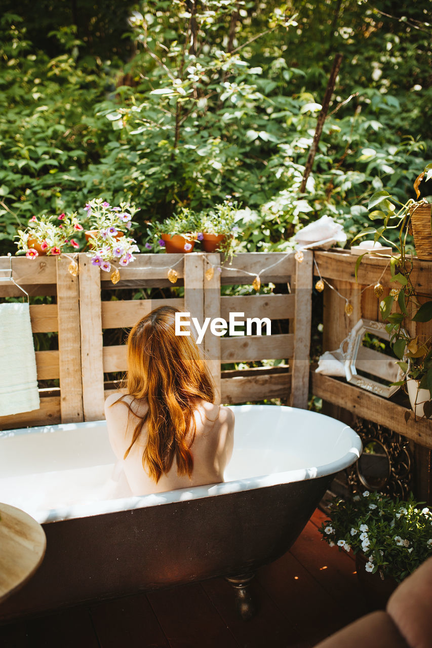 Back view of faceless topless thoughtful redhead female sitting in bathtub near wooden fence on patio during skin care routine on summer day