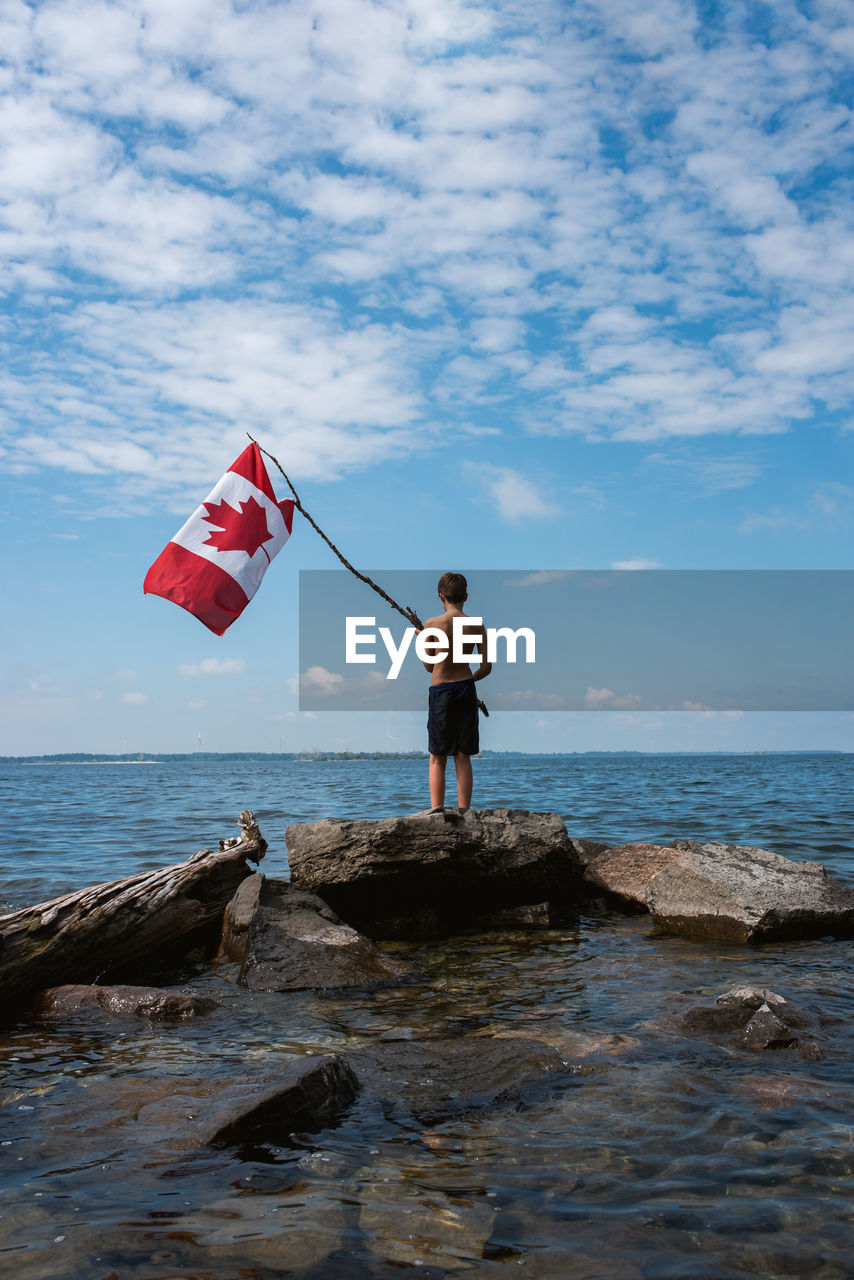 Boy holding canada flag in the air on shore of a lake on a summer day.