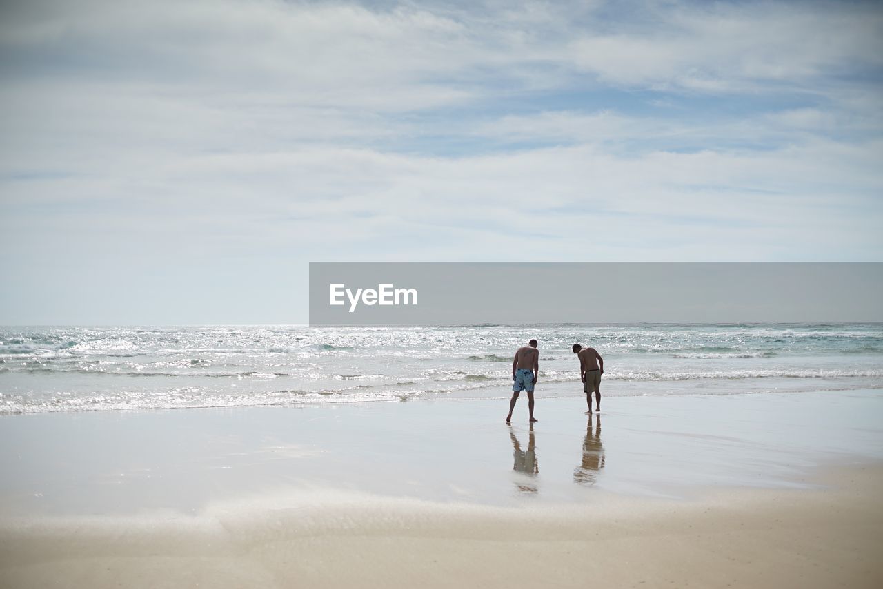 Men standing on wet shore at beach against sky