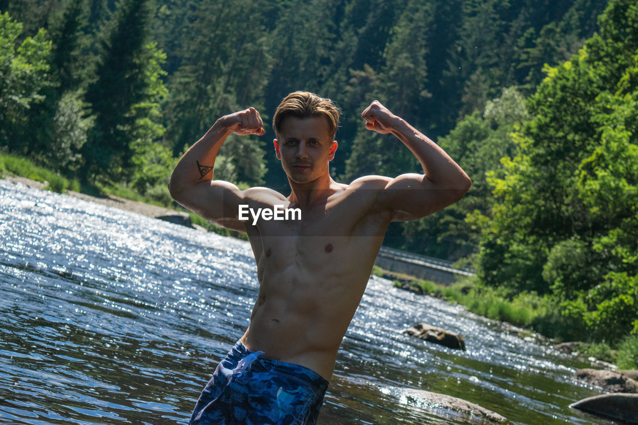 Portrait of shirtless man flexing muscles while standing by river in forest