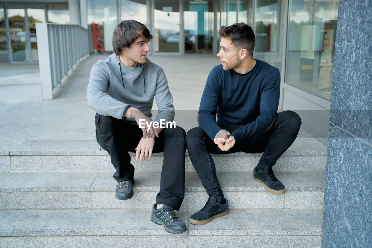 Front view of two young men talking sitting on a city staircase and
