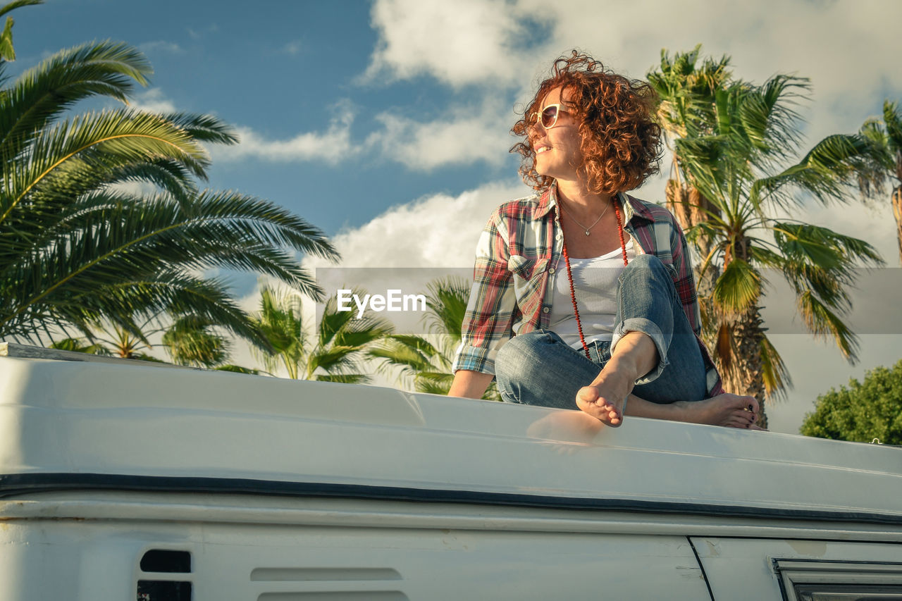 Low angle view of woman sitting on travel trailer against palm trees