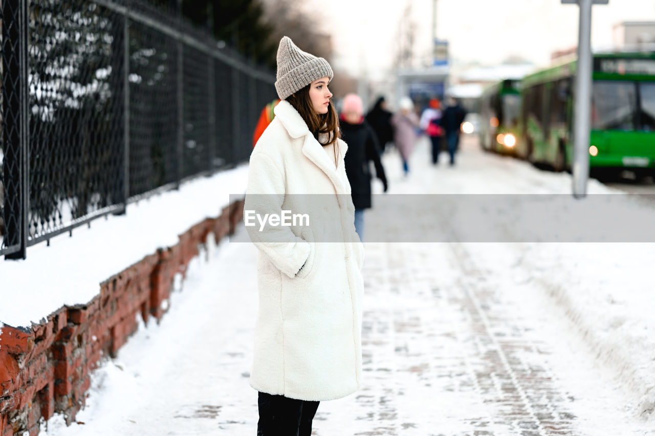 Girl in a fur coat walks along the road traffic of cars. the girl has a knitted hat on her head