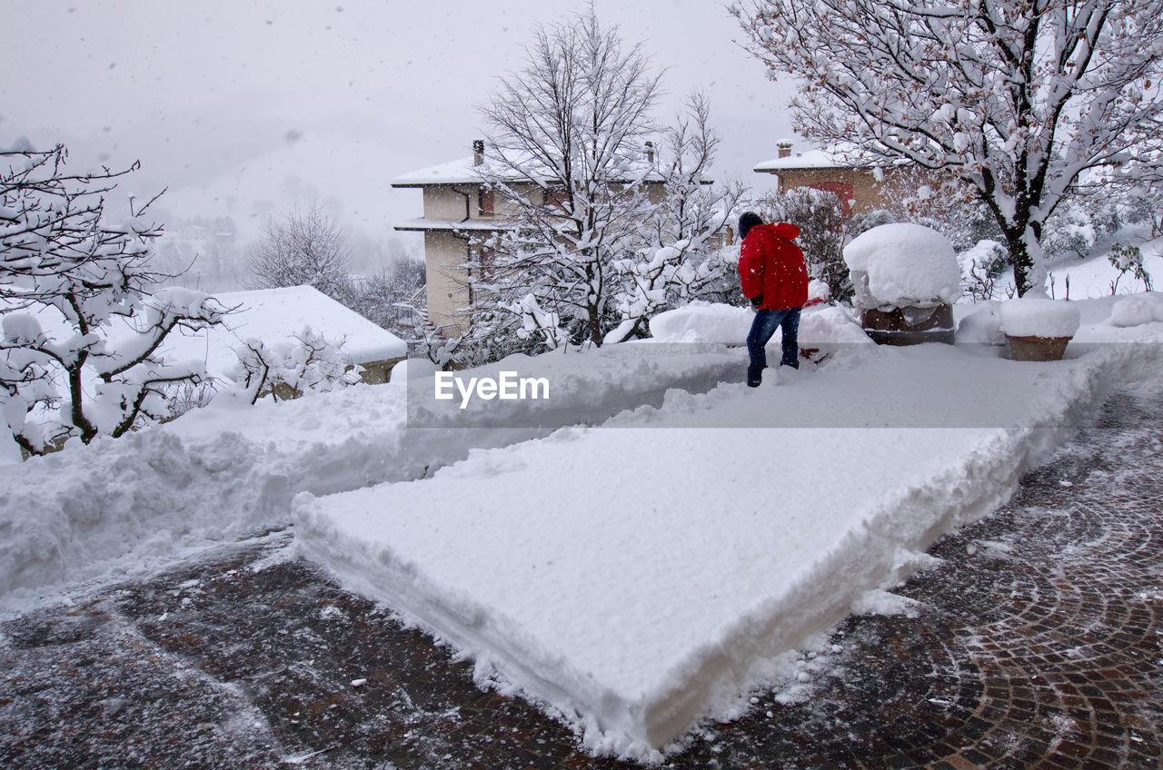 REAR VIEW OF MAN WALKING ON SNOW AGAINST BUILDINGS