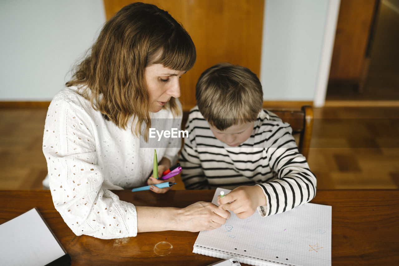 High angle view of mother teaching son with down syndrome to draw in book at table