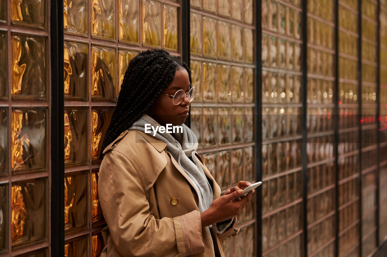 Confident black female with braids in casual outfit looking away and browsing smartphone while standing near glass wall