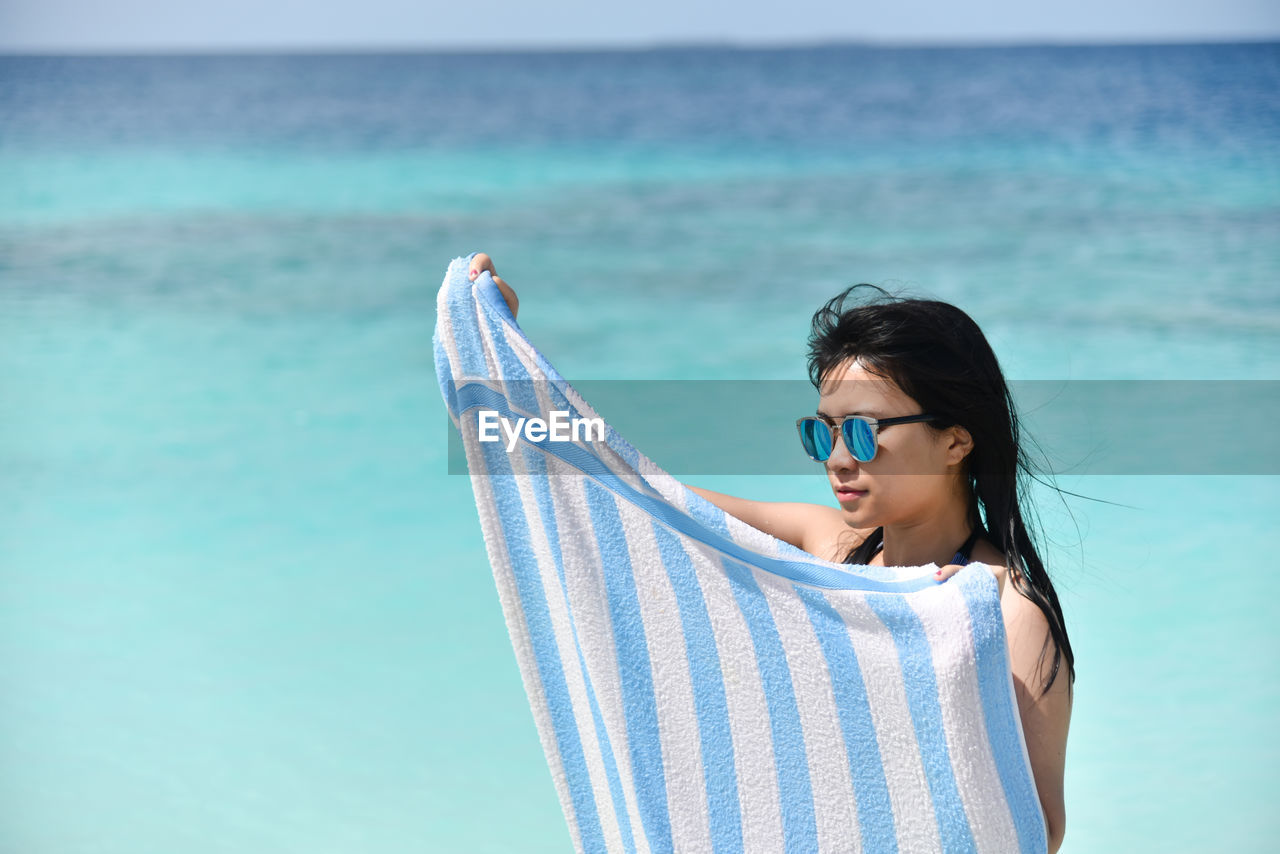 Young woman looking away at sea while holding towel against sky