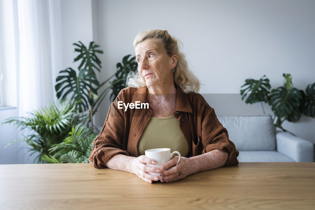 Sad elderly woman with mug sitting at home