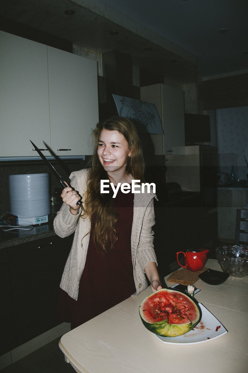 Smiling woman looking at knife while standing by watermelon at home