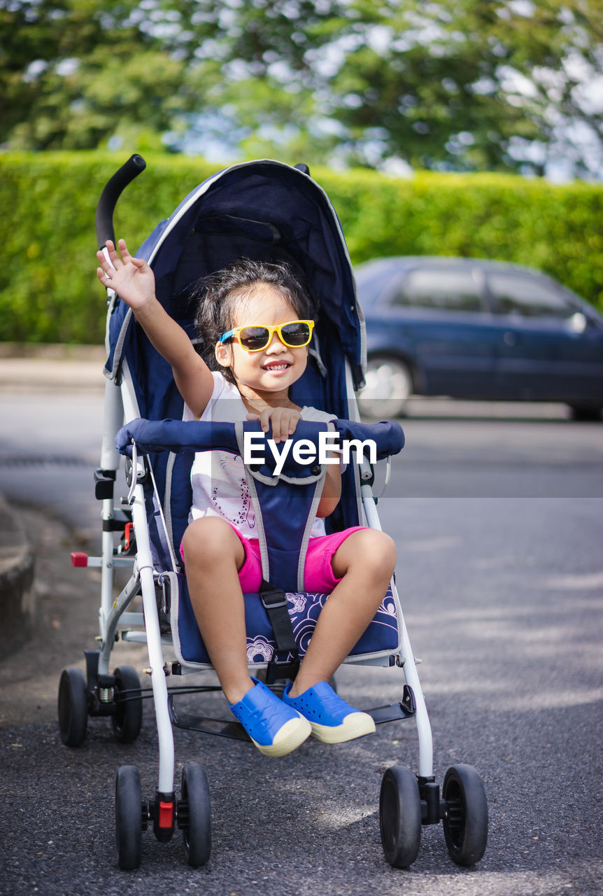 Full length of girl waving hand while sitting on baby stroller at street