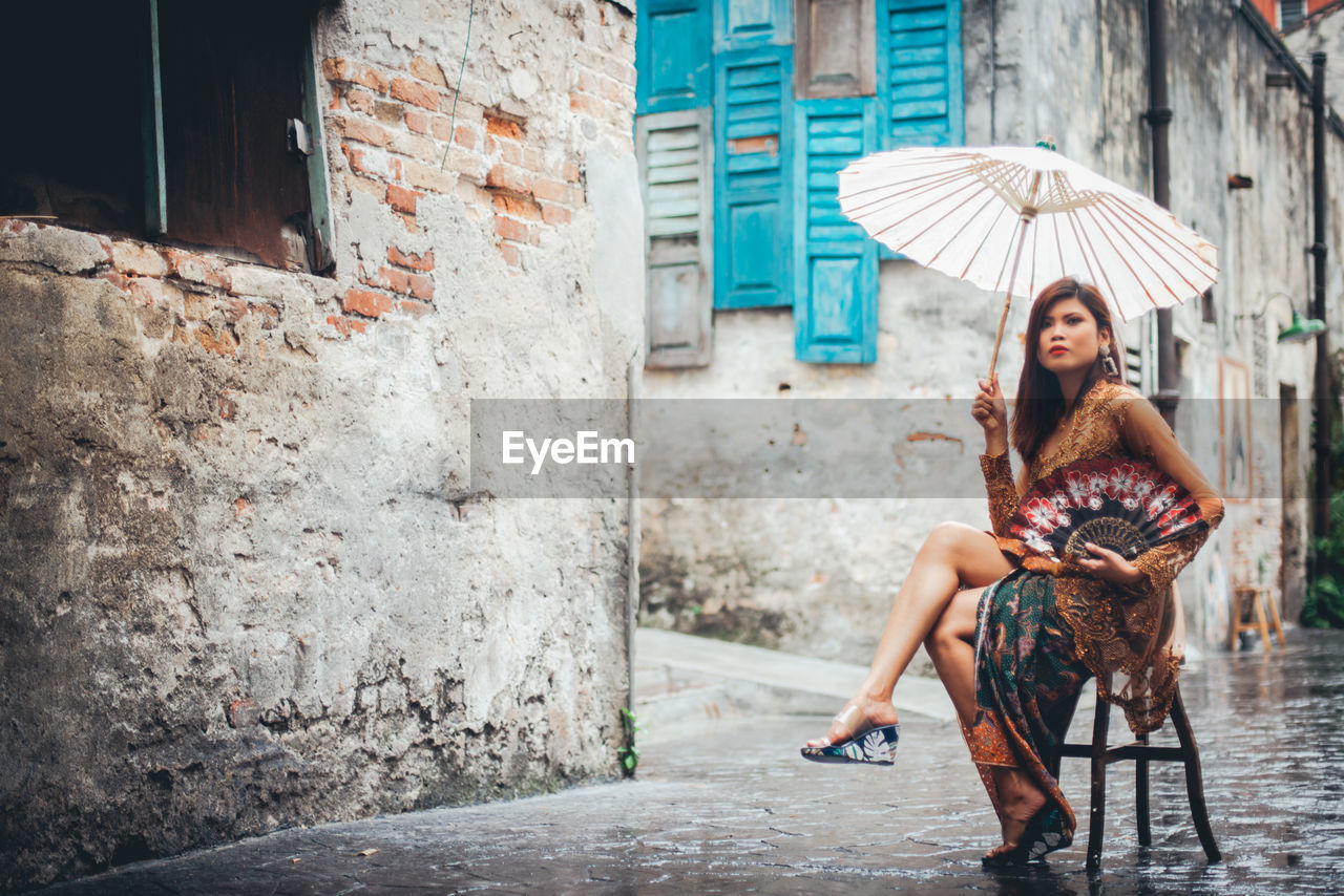 Full length of beautiful woman with umbrella sitting on stool in old town