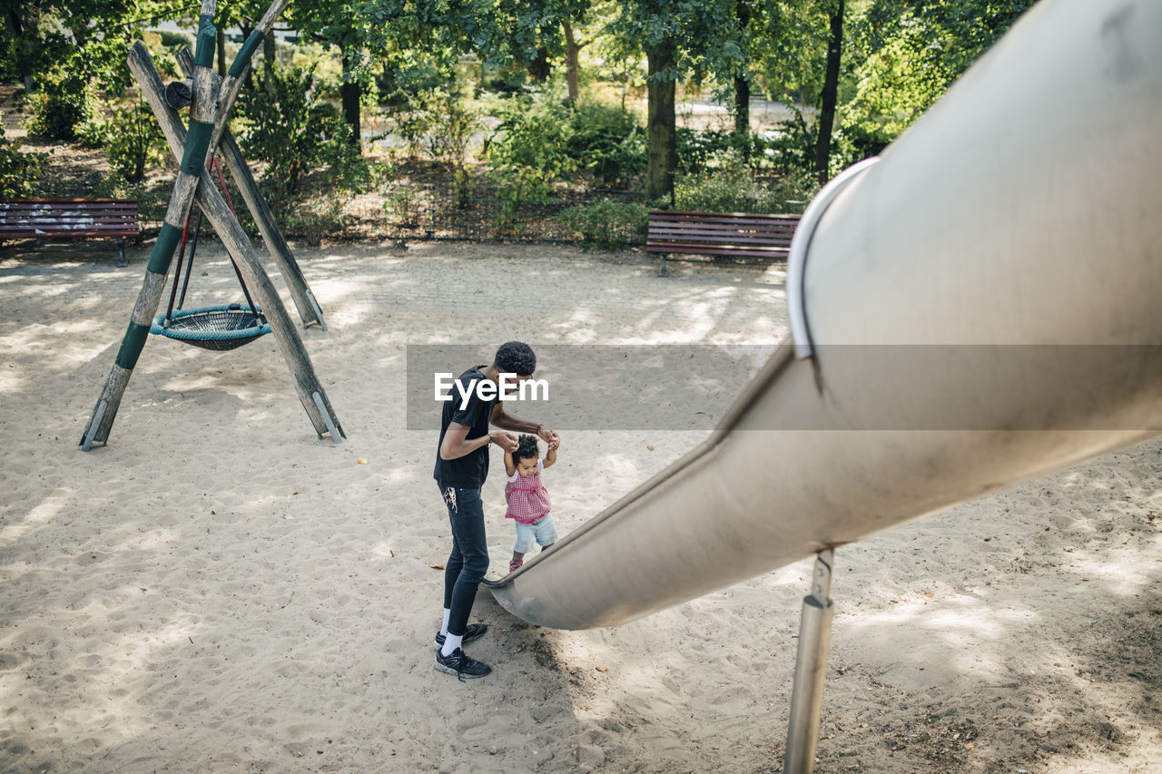 Father holding hands of daughter walking on slide at playground