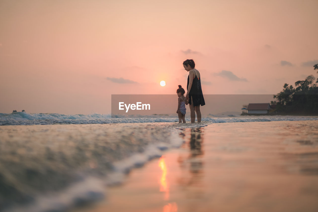 Mother with daughter standing at beach against sky during sunset